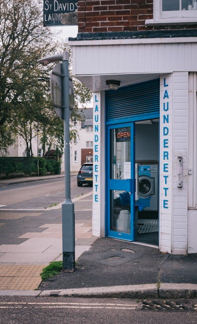Exterior view of a small laundrette on South Lambeth Road in Stockwell, showing a white wooden facade with blue vertical signage reading 'LAUNDERETTE' on both sides of a glass door. The door is partia